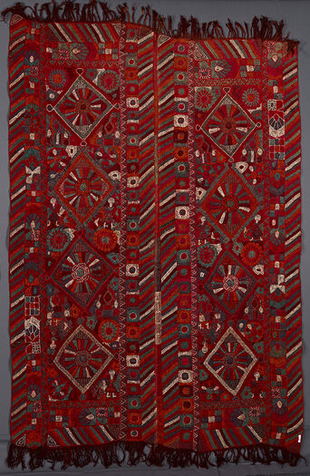 (UEM49205) The blanket has double vertical borders.&amp;#160; The characteristic multi coloured diagonal lines pattern are on all of the four vertical edges of the two panels. Each panel has four diamonds in which there is a large flower or a wheel-like motif with rays. There are girls surrounding two of the diamonds in two of their corners and in the other two diamonds in there stylised mudhifs (local guest houses ) in two of the corners. The embroidery is somewhat crude.&amp;#160;