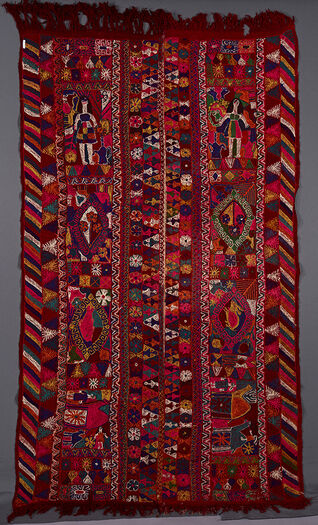 (UEM49208) This blanket has&amp;#160;double borders. Only the outer edges of the two panels have the characteristic multi coloured diagonal lines pattern.&amp;#160;The first&amp;#160;vertical panel consist of a chequeredboard of flowers, a girl with long hair wearing a short dress. In centre of the panel is a diamond with flowers, a diamond with a rabbit and two pomegranates, two long legged camels,&amp;#160;one which has a rider and chequered board of flowers.&amp;#160;
&amp;#160;
The&amp;#160;second vertical panel has a chequered board of flowers, a crowned female figure surrounded by animals and a coffee pot. In the centre of the panel there is a diamond with flowers surrounded by animals, a diamond with a fish surrounded by animals; a big and a small camel with a man holding one of them; chequeredboard of flowers.&amp;#160;&amp;#160;
The&amp;#160;colours are more garish and embroidery rougher than on the other blankets.&amp;#160;
&amp;#160;
The central area of each panel is divided into four sections which are further separated into panels of a floral motif. In the centre are human figures (female figures with short dresses), houses, camels,riders, fish, rabbits, scorpions, birds and multi-petalled flowers as well as four-petalled ones and a pomegranate motif. To be checked and completed.&amp;#160;