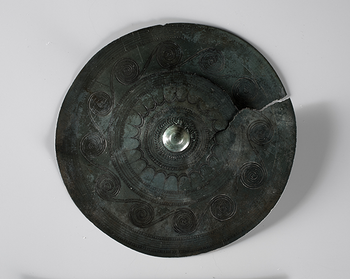 The belt plate from Vestre Evang on Toten in the Inland is one of the finest Bronze Age jewellery we have in the collection. It was found in a large burial mound around 1870. Probably the buried was a woman, who lived once between 1500 and 1300 BC. The belt plate is approx. 13 cm in diameter, and on the back, there is a small loop. The plate is decorated with spirals and sun symbols. The fine and precise lines show that the jewellery was made by a master blacksmith. Belt plates can be compared to today&#39;s National costume jewellery, and were worn in the belt on the women&#39;s suit. In 1971, almost 3300 years later, the belt plate from Evang was recreated as a jewellery needle for the Toten traditional costume. C5573.
