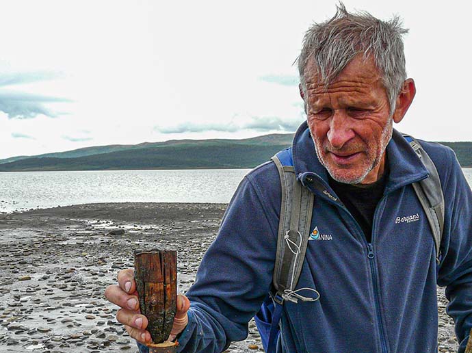 Mann holder en del av en flere tusen år gammel trestaur i hånda. I bakgrunnen ligger et fjellvann.